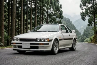 Toyota AE86 Trueno Corolla blanche sur une route de montagne japonaise entourée de forêt de cèdres, vue trois quarts avant
