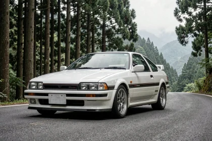 Toyota AE86 Trueno Corolla blanche sur une route de montagne japonaise entourée de forêt de cèdres, vue trois quarts avant